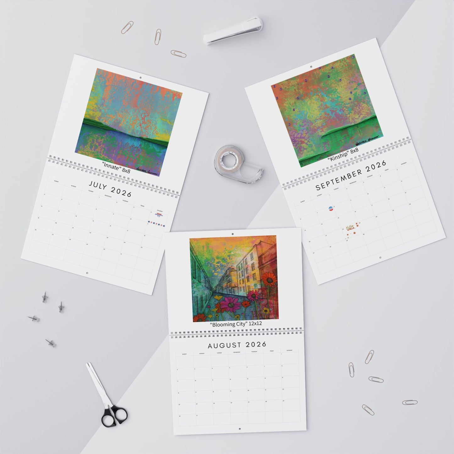 Three calendar pages showing colorful artwork on a light gray surface with stationery items.