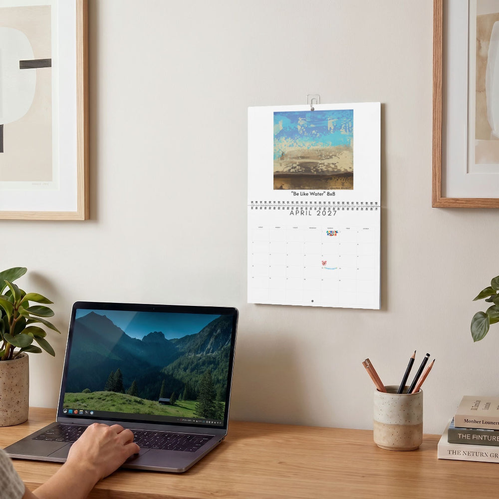 A wall-mounted calendar hangs on a beige wall above a wooden desk. A person's hand is visible using a laptop on the desk, with framed wall art, a plant, a pencil cup, and stacked books in the room.
