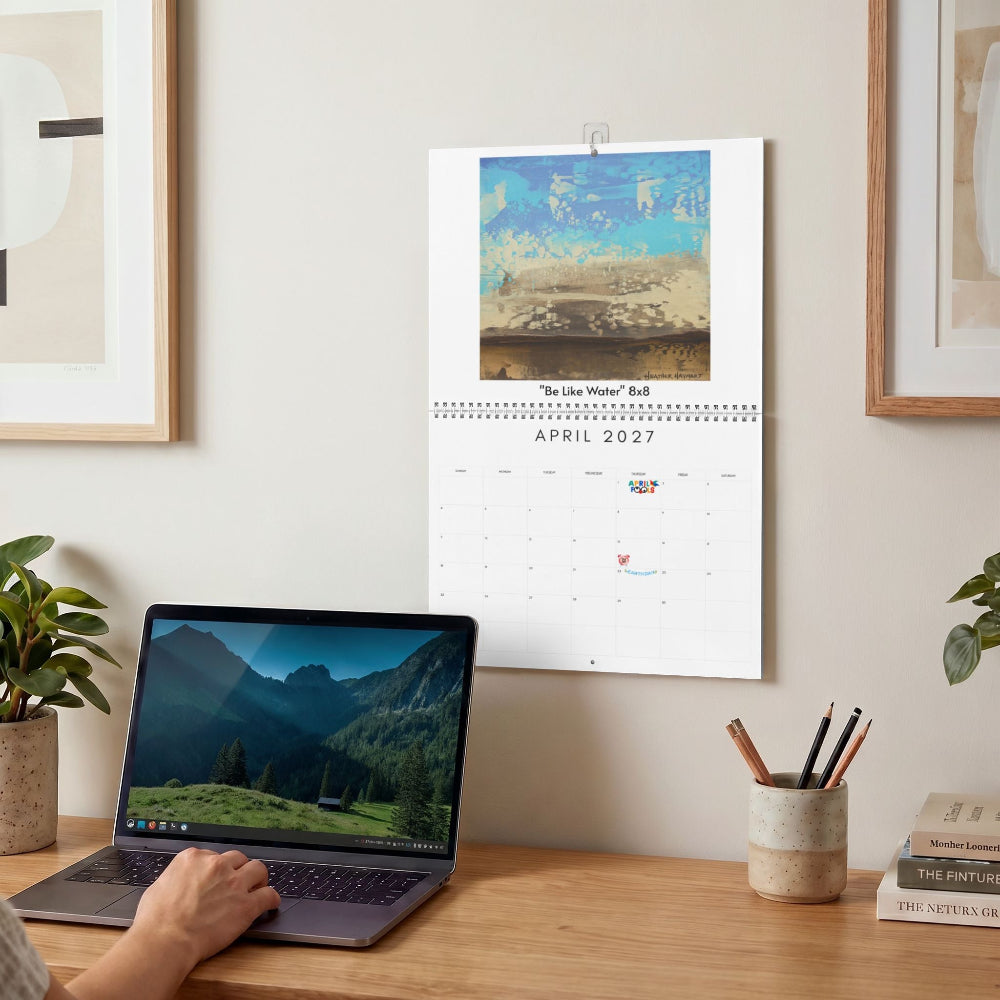 A wall-mounted 2027 calendar hangs on a beige wall above a wooden desk. The calendar features a landscape photograph at the top and a printed April 2027 page below.