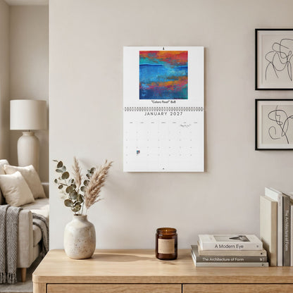 A January 2027 wall calendar featuring an abstract colorful painting, hanging on a beige wall in a styled living room setting with a wooden console table, books, and a vase.
