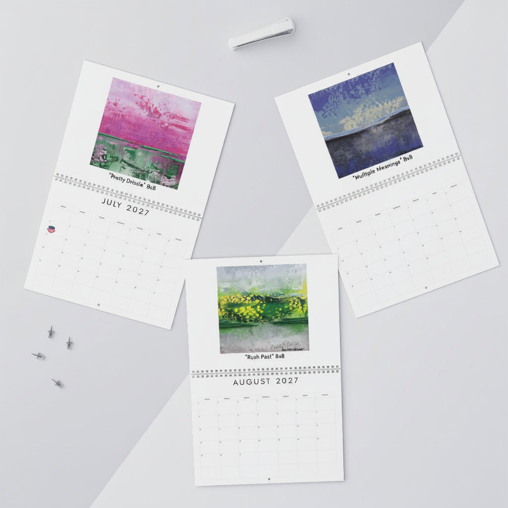 Three printed monthly calendar pages with watercolor landscape artwork are arranged on a light gray surface with small desk props including  and a pen.