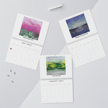 Three printed monthly calendar pages with watercolor landscape artwork are arranged on a light gray surface with small desk props including  and a pen.