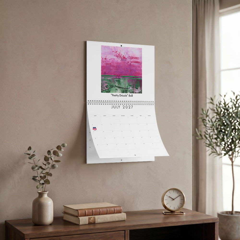 A wall-mounted calendar hanging on a beige wall in a styled interior. The calendar page for July 2027 is open, showing a colorful abstract artwork at the top and a grid below.