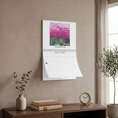 A wall-mounted calendar hanging on a beige wall in a styled interior. The calendar page for July 2027 is open, showing a colorful abstract artwork at the top and a grid below.