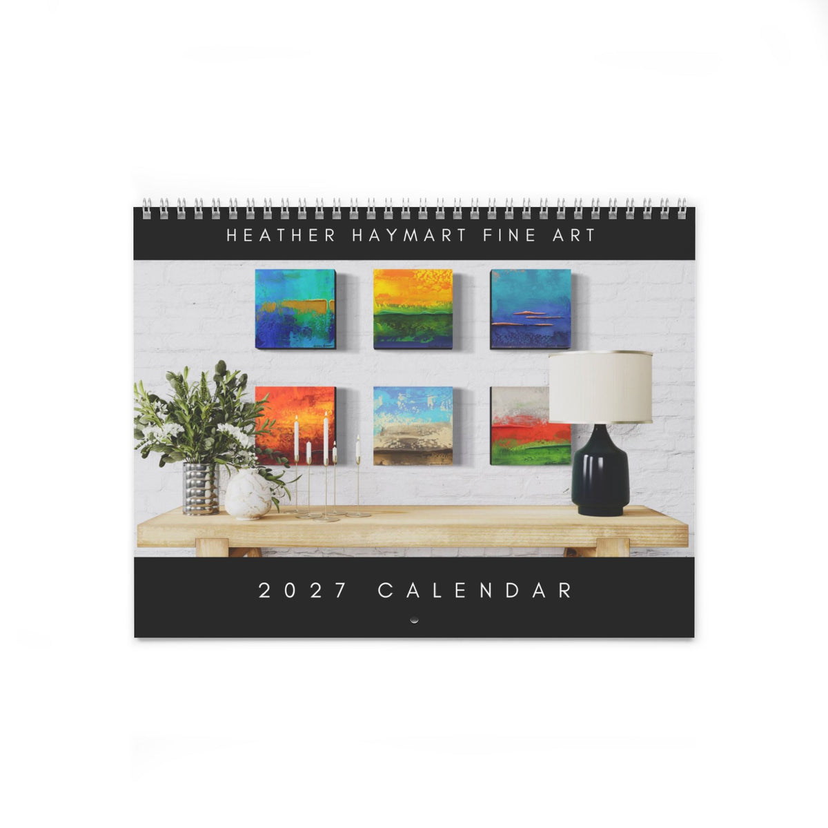 A spiral-bound 2027 calendar featuring a photograph of six colorful square paintings displayed on a white brick wall above a wooden console table with a vase of greenery and a black table lamp. The top and bottom of the calendar have black bands with white text reading 'HEATHER HAYMART FINE ART' and '2027 CALENDAR'.