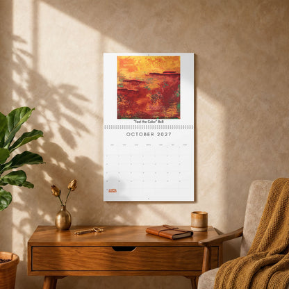 A vertical wall calendar page hanging on a beige wall in a styled room, featuring an abstract painting in warm orange, red, and yellow tones with the text 'Feel The Color' and 'OCTOBER 2027'. A wooden side table, plant, vase, and armchair with a throw blanket are visible below.
