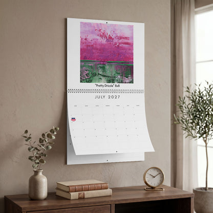 A wall-mounted calendar page for July 2027 hangs on a beige wall in a styled room. The top portion of the calendar features an abstract painting with pink, purple, and green tones.