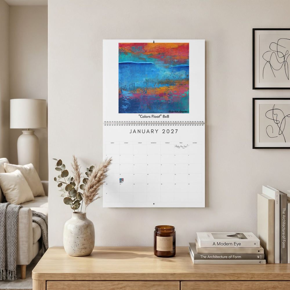 A wall-mounted calendar hanging in a styled living room. The calendar page shows the month January 2027 beneath an abstract painting with blue, orange, red, and magenta tones.