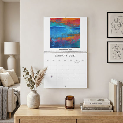 A wall-mounted calendar hanging in a styled living room. The calendar page shows the month January 2027 beneath an abstract painting with blue, orange, red, and magenta tones.