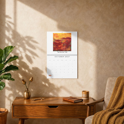 A wall-mounted calendar page with an abstract painting in warm orange, red, and yellow tones, displayed on a beige textured wall above a wooden side table in a styled room with a chair, plant, and sunlight shadows.