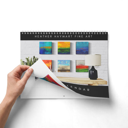 A spiral-bound wall calendar featuring Heather Haymart Fine Art prints, shown with a hand lifting one page to reveal the blank grid layout underneath. The cover displays a modern interior scene with colorful abstract paintings, a lamp, and a wooden table.