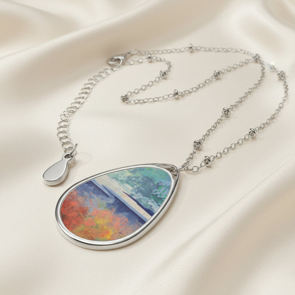 An art necklace with an ellipse-shaped pendant featuring a blend of colors including blue, red, and green, attached to a silver aluminum print surface on a chain.
