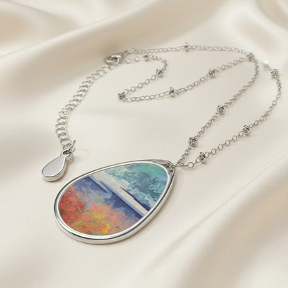 An art necklace with an ellipse-shaped pendant featuring a blend of colors including blue, red, and green, attached to a silver aluminum print surface on a chain.