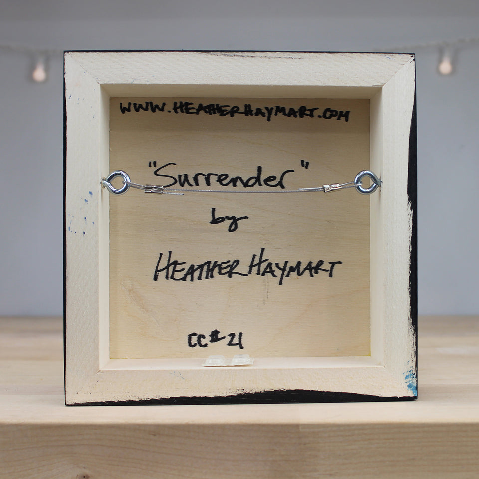 Small wooden frame with a metal bracelet labeled 'Surrender' by Heather Haymart on a neutral background.