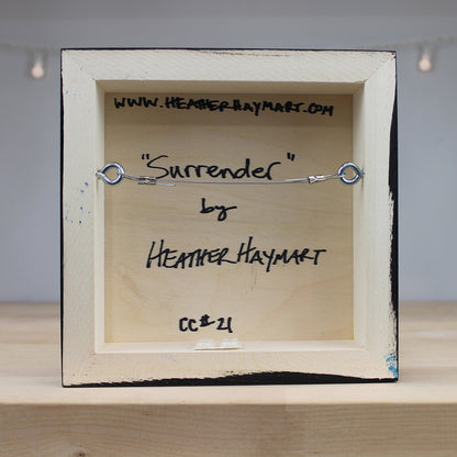 Small wooden frame with a metal bracelet labeled 'Surrender' by Heather Haymart on a neutral background.